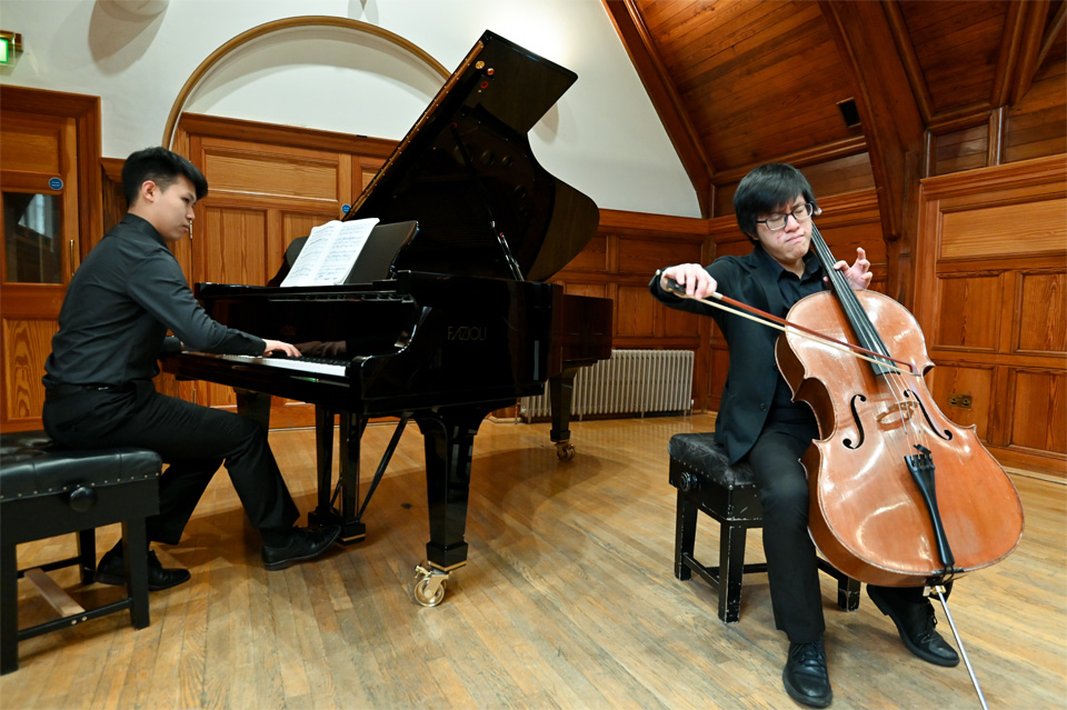 An Asian male student, playing the cello, with another student accompanying him on the piano, in a wooden room.